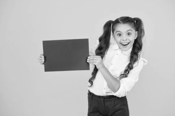 Skolan är en lycklig plats. Glad liten barn håller tomt rosa papper på gul bakgrund. Liten flicka glad leende med tom affisch. Det kommer att göra dig lycklig, kopiera utrymme — Stockfoto