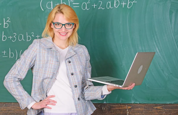 Woman teacher wear eyeglasses holds laptop surfing internet. Basic school education. Teacher elegant lady with modern laptop surfing internet chalkboard background. Distance education concept