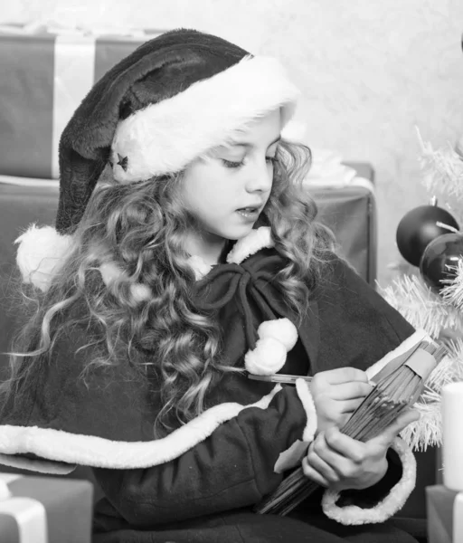 子執筆手紙サンタ クロース。親愛なるサンタ。女の子かわいい子は、クリスマス ツリーの近くに紙とペンを保持します。奇跡を信じています。サンタさんの手紙。しい物のリスト。子供サンタ帽子は、クリスマス ・ イヴをお楽しみください。 — ストック写真