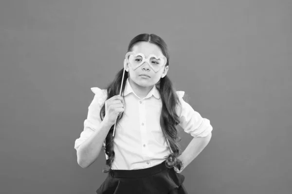 Concepto de fiesta escolar. Mirada inteligente niño a través de gafas de utilería cabina de fotos. Día de San Valentín. Símbolo de amor. Pequeña colegiala inteligente sobre fondo naranja. Niño pequeño con gafas de ojo estilo inteligente — Foto de Stock