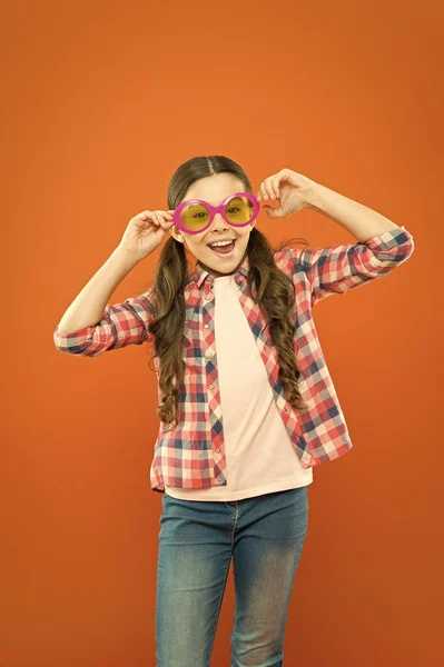 Rimanere concentrati con gli occhiali. Ragazzino sorridente con occhiali da festa fantasia su sfondo arancione. Ragazza divertente che indossa occhiali da sole con filtro a colori. Bambino piccolo cercando felice attraverso gli occhiali di moda — Foto Stock