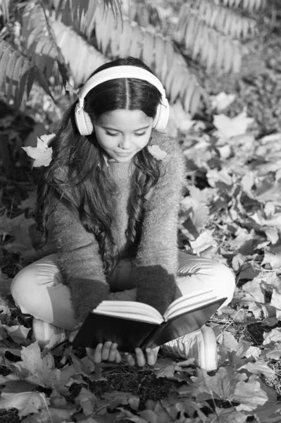 오디오 학습의 기회입니다. 스테레오 이어폰을 끼고 가을 나뭇잎에 앉아 있는 귀여운아이. 어린 아이는 오디오 학습을 좋아 합니다. 어린 소녀는 헤드폰으로 오디오 책을 듣는다. 교훈이 되는 음향 자료 — 스톡 사진