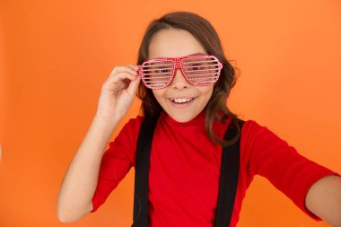 Selfie çekiyordum. Eğlence için parti gözlükleri. Çocuk modası konsepti. Gözlüklere odaklanıyorum. Havalı görünümlü turuncu arka plan. Güzel ve şık bir çocuk. Gülümseyen küçük kız okul üniforması