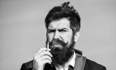 Sakallı ve bıyıklı bir adam sigara tutuyor. Sakallı hipster sigara mavi gökyüzü arka plan sigara. Sigaralı adam nikotin etkisinin tadını çıkarıyor. Sigara içmenin zevki. Rahatlatıcı ama zararlı alışkanlık