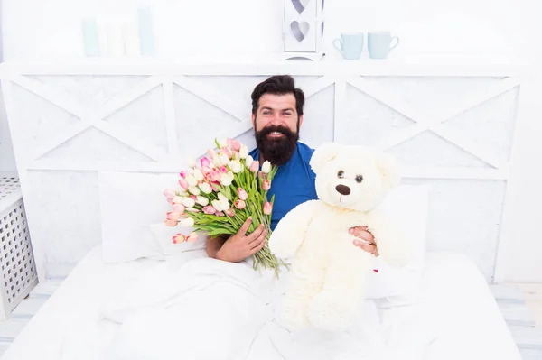 Happy moments. good morning surprise. happiness about present. cheerful bearded man in bed. happy birthday gift. spring fresh tulip and bear toy. love valentines day. womens day. flowers for march 8