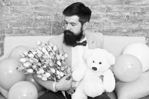 Romantic gift. Romantic man with flowers and teddy bear sit on couch with air balloons waiting girlfriend. Macho ready romantic date. Man wear tuxedo bow tie hold bouquet. She deserve all best — Stock Photo, Image