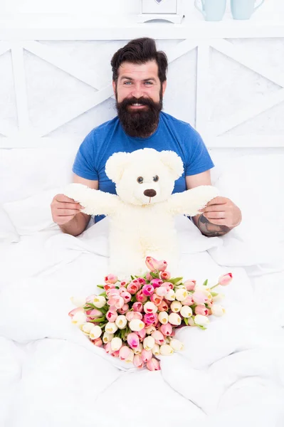 Férias de aniversário. Faça um conceito surpresa. Presente para o cônjuge. Urso de peluche. Flores frescas. Um hipster barbudo na cama. Presente do Dia dos Namorados. Homem segurar tulipas buquê relaxante na cama. Serviço de entrega de flores — Fotografia de Stock