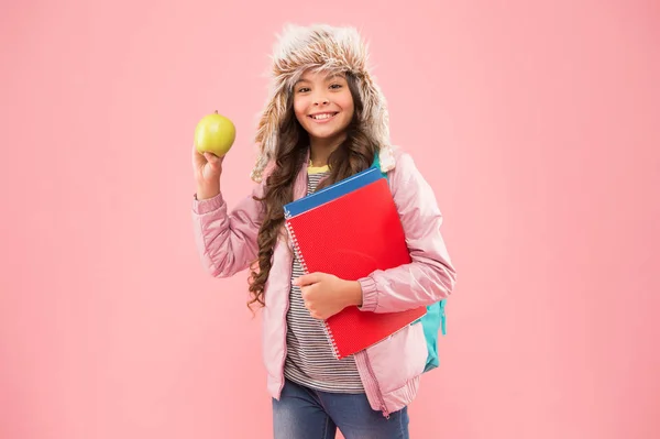 Έχει υπέροχη γεύση. Μαθήτρια με βιβλία μετά το μάθημα. Σχολικές διακοπές. μικρό χαρούμενο κορίτσι με καπέλο που τρώει μήλο. χειμερινές διακοπές και διακοπές. έφηβος παιδί ροζ φόντο. την καθημερινή ζωή των μαθητών. Σύγχρονη εκπαίδευση — Φωτογραφία Αρχείου