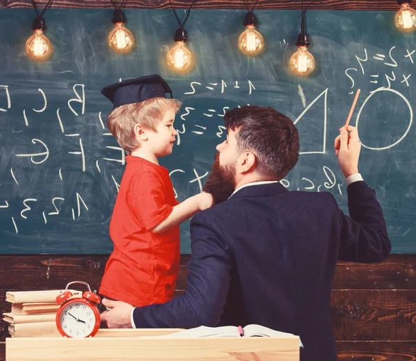 Teacher with beard, father teaches little son in classroom, chalkboard on background. Instructive conversation concept. Child in graduate cap listening teacher, chalkboard on background, rear view