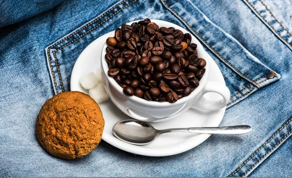 Tasse mit Kaffeebohnen, raffiniertem Zucker und Löffel auf Teller, Jeanshintergrund. Kaffeepause-Konzept. Becher voller Kaffeebohnen und Haferkeks auf Jeans — Stockfoto