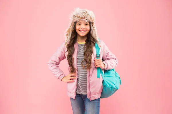 Pod koniec roku szkolnego. Żadnych więcej egzaminów. Mała szczęśliwa dziewczynka wraca do szkoły. ferie zimowe i wakacje. dziecko ciepłe ubrania różowe tło. szczęśliwy kapelusz klapki dla dzieci. uczennica z plecakiem idź do domu — Zdjęcie stockowe