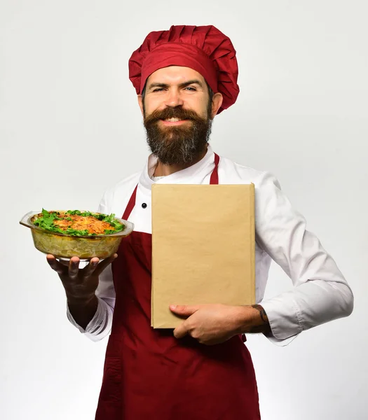 Cook with happy face in burgundy uniform holds baked dish — Stock Photo, Image