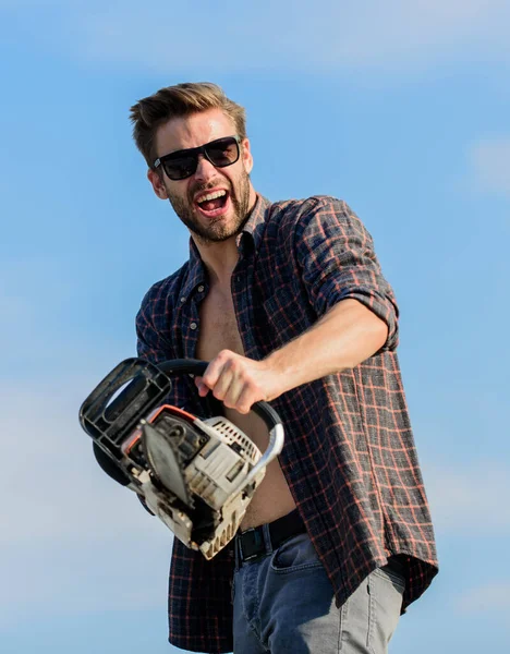 Handsome man with chainsaw blue sky background. Gardener lumberjack equipment. Lumberjack with chainsaw in his hands. Masculinity concept. Sharp blade. Dangerous job. Feeling manly. Powerful chainsaw