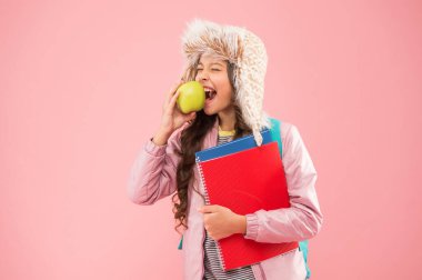 Hayatı daha iyi hale getirmek. Günlük öğrenci hayatı. Modern eğitim. Ders sonrası kitapları olan liseli kız. Okul tatili. Küçük mutlu kız kulaklı şapka elma yer. Kış tatili ve tatil. genç pembe arka plan.