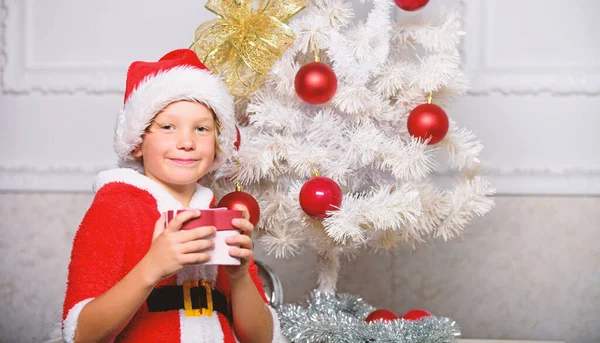 Хлопчик, одягнений як Санта з червоним капелюхом, тримає подарункову коробку біля ялинки. Різдвяний костюм Санти Клауса для дитини. Різдвяна вечірка з костюмом Санти. Ідеї новорічної ялинки для дітей — стокове фото