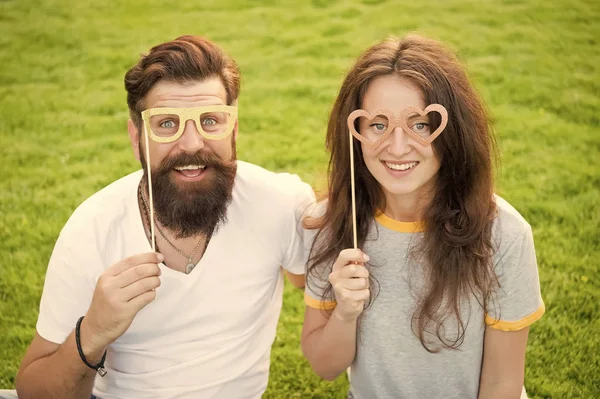 Fun Valentines props. Bearded man and sexy girl smiling with fancy party props on green grass. Funny couple holding heart shaped photobooth props on stick. Happy family celebrating with glasses props