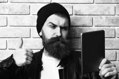 Sakallı, beyaz tenli hippi bej tuğla duvar stüdyosunun arka planında tablet taşıyor.