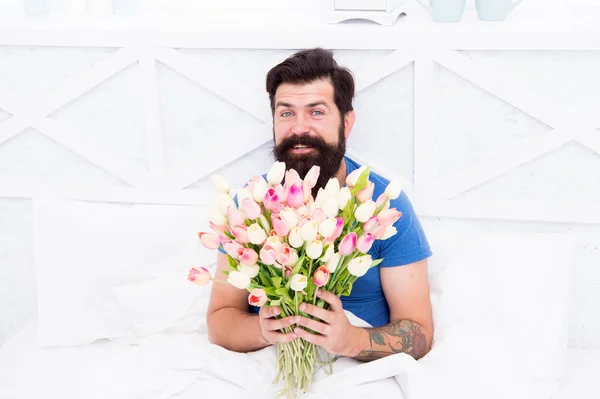 Man hold tulips bouquet while relaxing in bed. Tender bloom. Fresh flowers surprise. Flowers delivery service. Birthday anniversary holiday. Gift for spouse. Bearded hipster in bed. Spring in bedroom