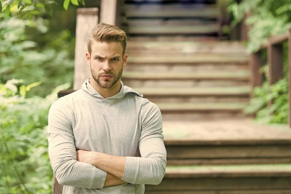 Muscular and confident. Muscular man on summer day. Athletic guy keeping muscular arms crossed at park stairs. Handsome sportsman or athlete showing muscular strength and power