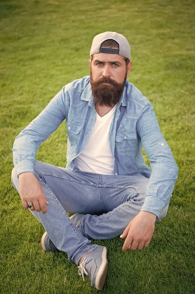 Obejmując estetykę sklep streetwear. Sexy mężczyzna siedzi na zielonej trawie. Brodaty człowieka w stylu casual denim. Kaukaski mężczyzna z wąsy i zarost. Brutalny mężczyzna z nieogoloną twarzą w kapelusza SnapBack — Zdjęcie stockowe