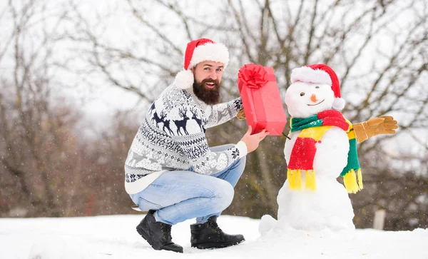 Winter games. Guy happy face snowy nature background. Hipster with beard hold gift box. Surprise concept. Winter activity. Winter vacation. Man made snowman. Man Santa hat having fun outdoors