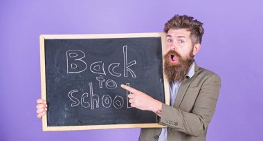 Teacher bearded man stands and holds blackboard with inscription back to school violet background. Continue your education with us. Teacher invites to continue studying. New semester in school clipart