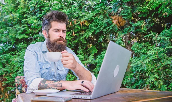 El trabajo y el juego le dan descanso y energía. Hombre barbudo haciendo su trabajo en línea. Hipster beber té y el uso de la computadora estación de trabajo al aire libre. Trabajar globalmente a distancia —  Fotos de Stock