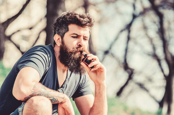 Хипстерский человек держит ваппинговый прибор. Бородатый жестокий мужчина курит электронную сигарету. мужчина курит электронную сигарету. Безопасность и зависимость от здоровья. Вдыхаю пар. Зрелый хипстер с бородой. Утренняя программа
