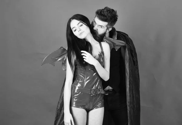 Couple in love play role game. Vampires victim concept. Man and woman dressed like vampire, demon, red background. Vampire in cloak behind sexy devil girl. Vampire bites female neck