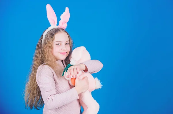 Cute and adorable. Bunny girl with cute toy on blue background. Child smiling play bunny toy. Happy childhood. Get in easter spirit. Bunny ears accessory. Lovely playful bunny child with long hair