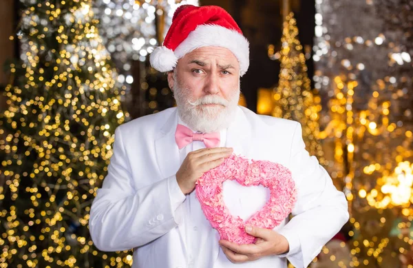 Esto es para ti. serio hombre de negocios corazón decorativo. le encantan las fiestas de Navidad. el invierno puede ser alegre. senior santa en esmoquin sobre fondo de luces de Navidad. guirnalda decoración de año nuevo. regalo con amor — Foto de Stock