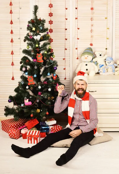 Guy in hat and scarf sits by Christmas tree. — Stock Photo, Image