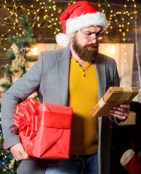 Man bearded hipster wear santa hat hold bunch of letters and gift box. Man bearded with eyeglasses reading post for santa. Spirit of winter miracle. Post for santa claus. Letter for santa claus