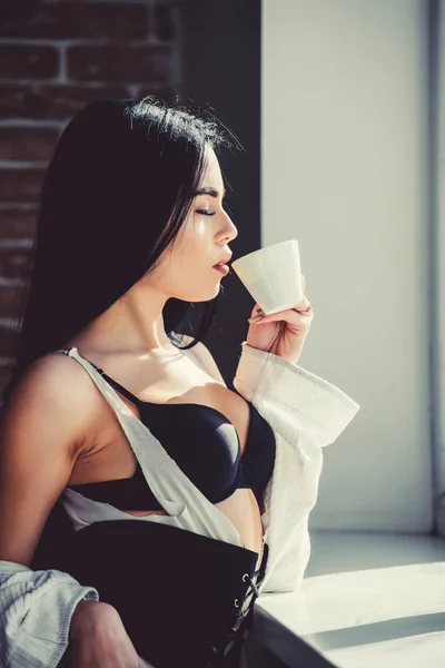 Наслаждаюсь утренним солнцем. Чувственная девушка сексуальная грудь пьет кофе возле подоконника. Привлекательное женское белье расслабляет дома. Страстный таинственный любовник. Идеальное время для кофе. Но сначала кофе. — стоковое фото