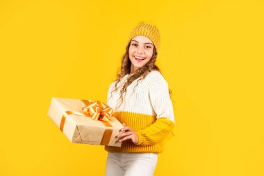Mutlu Noeller ve mutlu tatiller. Mutlu kış tatilleri. Neşeli küçük kız örgü şapka ve kazak örüyor. Çocuk hediye kutusunu sarı arka planda tutuyor. Xmas hediye alışverişi. Noel 'in aydınlık olsun.