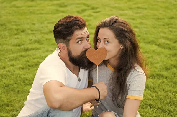 Given his heart to her. Sensual couple in love with prop heart on green grass. Sexy woman and bearded man holding valentines heart on stick. One heart two souls