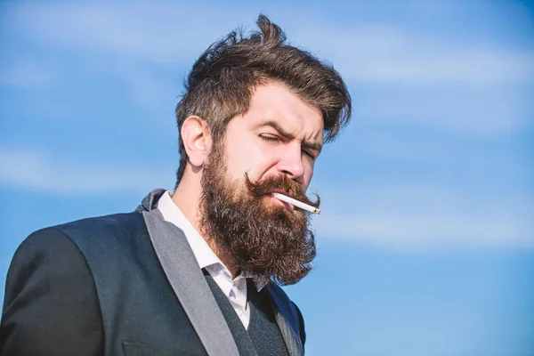 Truth about smoking pleasure and nicotine addiction. Bearded hipster smoking cigarette blue sky background. Guy with cigarette enjoy nicotine influence. Man with beard and mustache hold cigarette
