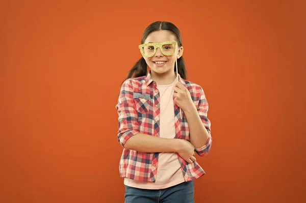 Περίεργο βλέμμα. Αξιολάτρευτο κορίτσι χαμογελά με το κόμμα βλέμμα σε πορτοκαλί φόντο. Χαριτωμένο μικρό παιδί που έχει χαρούμενο βλέμμα μέσα από τα γυαλιά. Μόδα εμφάνιση του μικρού μοντέλου μόδας — Φωτογραφία Αρχείου