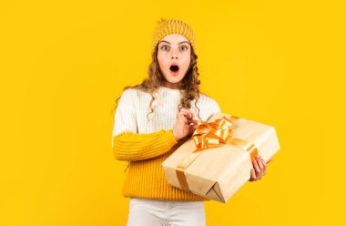 Çocuk hediye kutusunu sarı arka planda tutuyor. Xmas hediye alışverişi. Noel 'in aydınlık olsun. Mutlu kış tatilleri. Şaşırmış küçük bir kız şapka ve kazak örmüş. Mutlu Noeller.