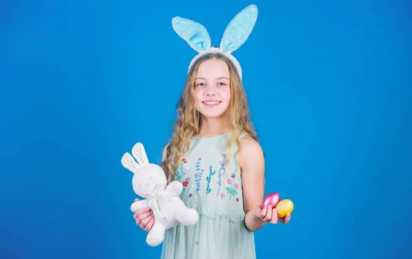 Glad påsk. Lycklig liten flicka klädd påsk bunny öron. Liten flicka i bunny pannband holding färgade ägg och leksak. Bedårande flicka barn med söt uppstoppad kanin leksak. Påskharen är symbol för påsk — Stockfoto