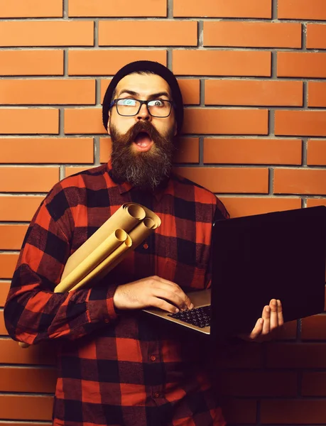 Rollos de papel artesanal en camisa a cuadros negro rojo con sombrero y gafas sobre fondo de estudio de pared de ladrillo marrón. Barbudo brutal caucásico hipster celebración de ordenador portátil y papel artesanal —  Fotos de Stock