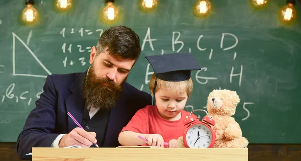 Miúdo ocupado a estudar na escola. Conceito individual de estudo. Professor em desgaste formal e aluno em papelão em sala de aula, quadro-negro em segundo plano. Professor, pai verificando lição de casa, ajuda a menino, filho — Fotografia de Stock