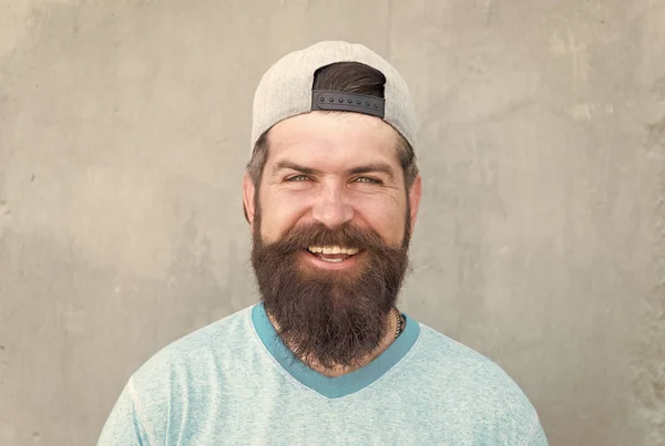 Keeping hair healthy. Happy hipster with shaped beard and styled hair wearing baseball cap. Bearded man smiling with unshaven face hair. Hairy guy with stylish beard and mustache hair on grey wall