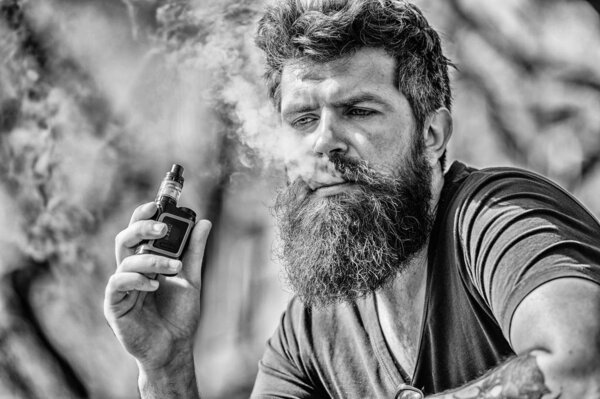 Человек с бородой выдыхает дым. Облака ароматизированного дыма. Бородатый человек курит вапе. Курение электронных сигарет. Концепция снятия стресса. Курящее устройство. Мужчина длинная борода расслаблена с привычкой курить

