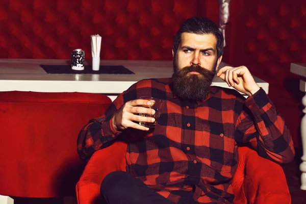 Γενειοφόρος hipster άνθρωπος κρατώντας ένα ποτήρι ουίσκι σε κόκκινη καρέκλα — Φωτογραφία Αρχείου