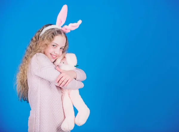 Bunny girl with cute toy on blue background. Child smiling play bunny toy. Happy childhood. Get in easter spirit. Bunny ears accessory. Lovely playful bunny child with long hair. Cute and adorable