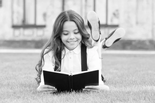 Grundbildung. Liebenswertes kleines Mädchen lernt lesen. Schuluniform für Schulmädchen auf dem Rasen mit Lieblingsbuch. Studienkonzept. Extraschulische Lektüre. Niedliches kleines Kind liest Buch im Freien — Stockfoto