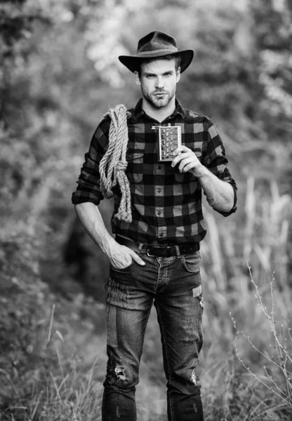 Cowboy ranch worker. Bourbon whiskey. Western culture. Man wearing hat hold rope and flask. Lasso tool American cowboy. Brutal cowboy drinking alcohol. Man handsome cowboy nature background — Stock Photo, Image