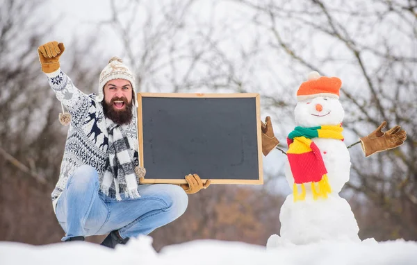 Téli ünnepek. Guy és hóember havas természet háttér. Téli esemény. Közelgő esemény. Egy szakállas férfi tartja a táblát a fénymásolóban. Téli bejelentés. Hipster kötött kalap és kesztyű mutatja tábla — Stock Fotó