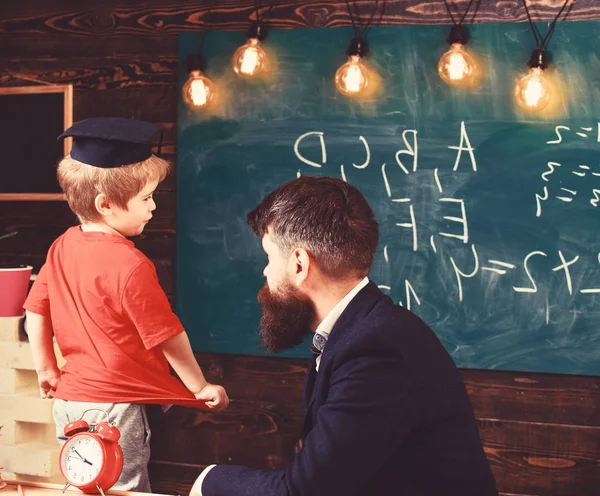 老师和孩子在教室里说话。牛津帽上的小男生在绿板前面回答这个问题。男成人坐在办公桌旁, 孩子站在他身旁. — 图库照片